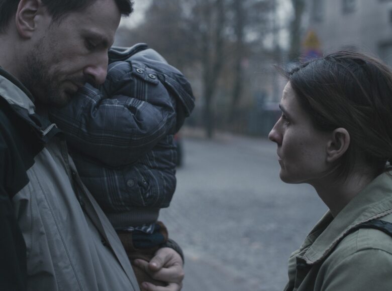 Kadr z filmu. Mężczyzna i kobieta stoją na ulicy przodem do siebie, mężczyzna trzyma na rękach dziecko. W tle szary miejski pejzaż.