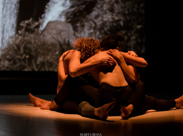 Spektakl teatralny. Trzy kobiety w obcisłych spodniach, z nagimi torsami i boso, siedzą na scenie w kręgu obejmując się. Za nimi scenografia przedstawiająca nocny, roślinny krajobraz.