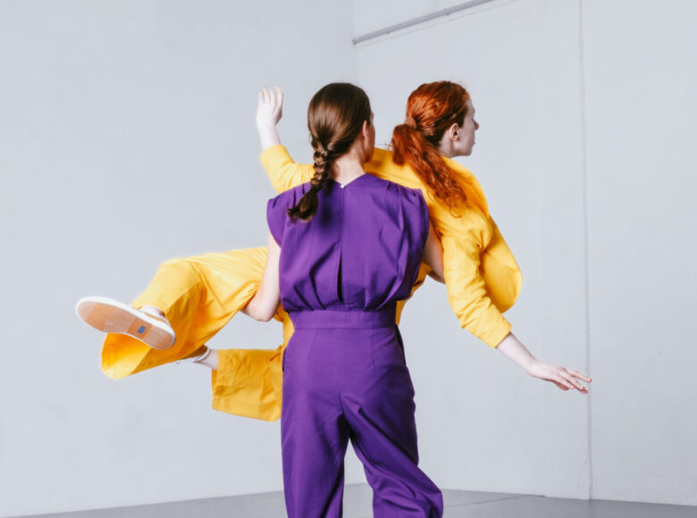 Kolorowa fotografia. Na jasnej sali prób dwie tańczące kobiety. Jedna z nich ubrana w fioletowy strój podnosi na rękach przed sobą drugą z nich ubraną na żółto. Fotografia jest dynamiczna.