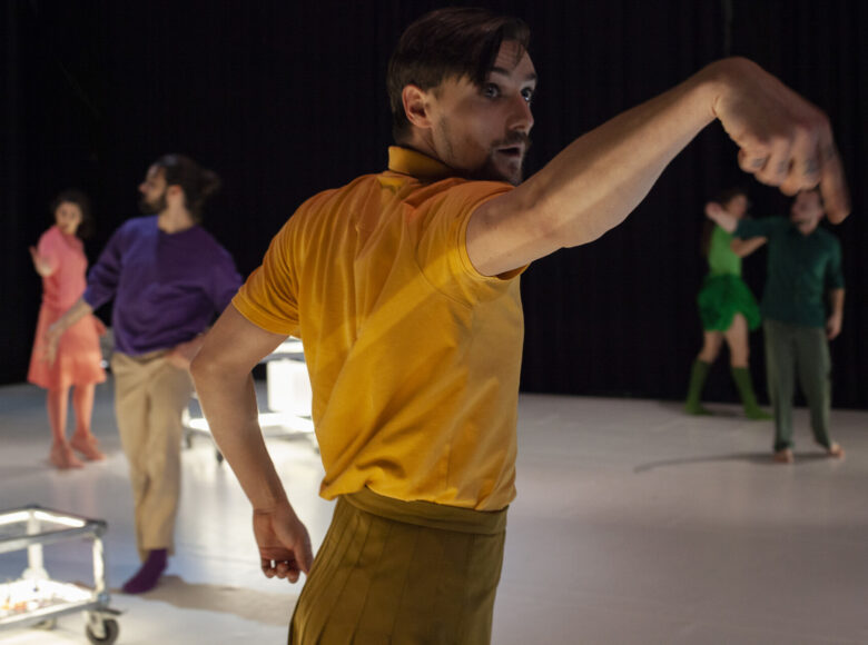 Kolorowa fotografia. Na pierwszym planie na scenie młody mężczyzna w żółtej koszulce stoi bokiem i dynamicznym gestem wyciąga do boku rękę. W tle na scenie 4 inne tańczące postaci.