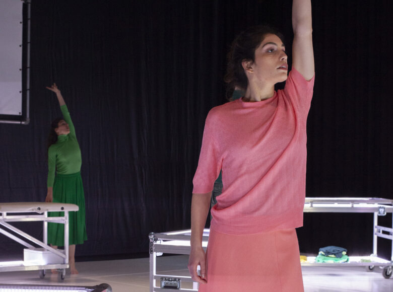 Kolorowa fotografia. Na pierwszym planie stoi młoda kobieta ubrana w różową sukienkę i wyciąga lewą rękę wyprostowaną do góry. Na drugim planie w tle sceny stoją 3 rozkładane łóżka na kółkach oraz kobieta w zielonej sukience, która też podnosi lewą rękę do góry.