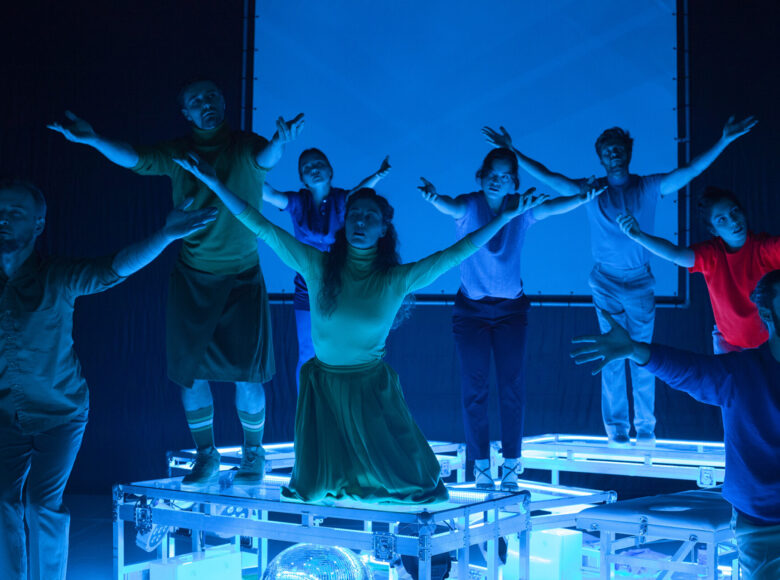 Kolorowa fotografia. Na scenie oświetlonej mocnym niebieskim światłem stoją podesty na kółkach. Na podestach stoi w tanecznych pozach kilka mężczyzn i kobiet. Wszyscy wyciągają przed siebie proste ręce w powitalnym geście.