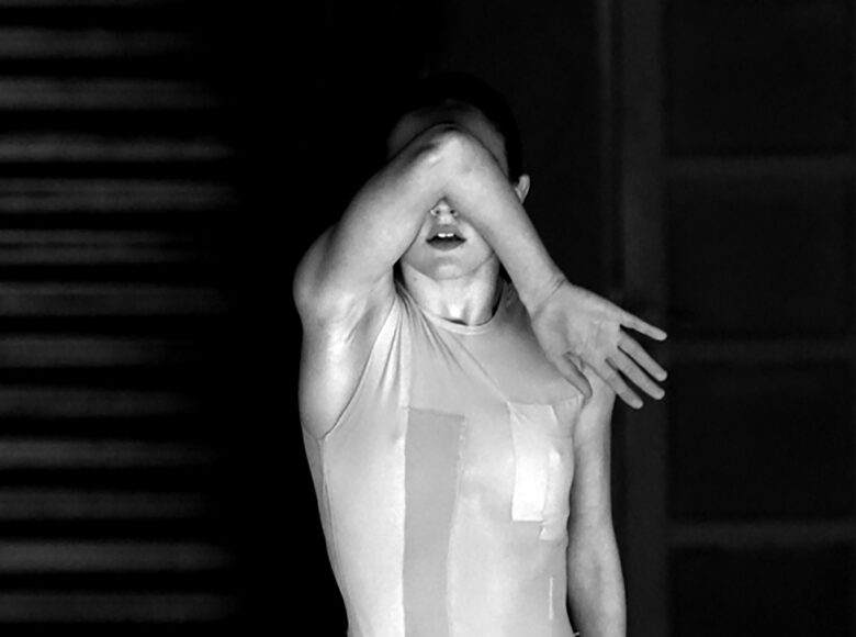 Czarno-biała fotografia. Na czarnym tle stoi młoda kobieta ubrana w jasną koszulkę bez rękawów, widać ją od pasa w górę. Łokciem prawej dłoni zasłania twarz, jakby raziło ją światło.