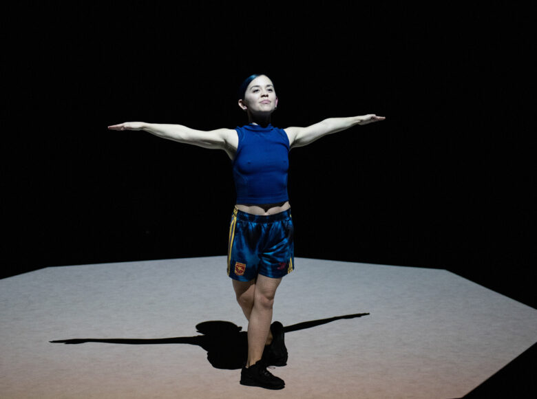 Kolorowa fotografia. Na środku ciemnej sceny stoi młoda drobna kobieta z ciemnymi włosami. Ubrana jest w krótkie sportowe spodenki i koszulkę bez rękawów. Stoi wyprostowana z rękami rozciągniętymi w bok, uśmiecha się i patrzy przed siebie.