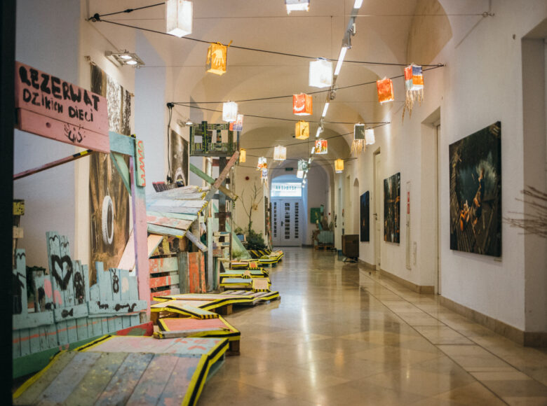 Zdjęcie korytarza galerii Sztuczki. W zadłuż ścian rozłożone są drewniane konstrukcje wykonane podczas zajęć w Rezerwacie Dzikich Dzieci. Do sufitu przyczepiona jest czarna lina z kolorowymi lampionami.