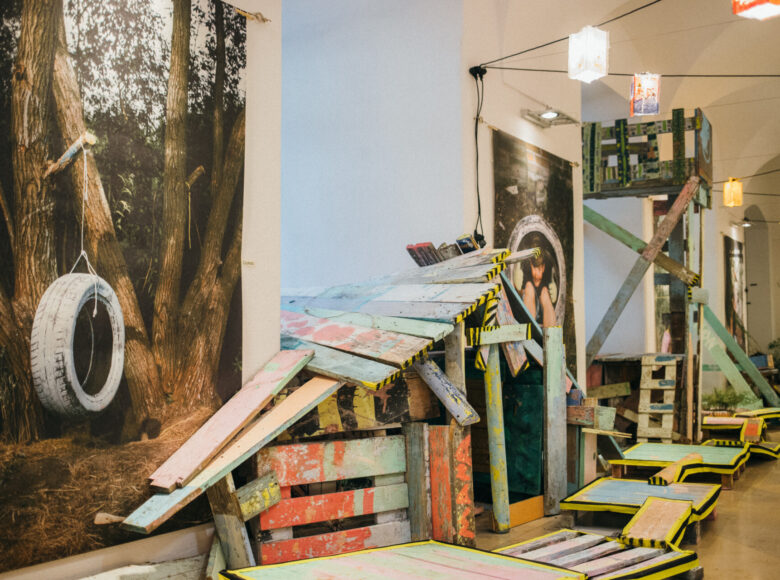 Przestrzeń Galerii Sztuczki. W pomieszczeniu znajdują się drewniane konstrukcje, pomalowane kolorową farbą, wykonane podczas zajęć w Rezerwacie Dzikich Dzieci. Na ścianach wiszą wielkoformatowe zdjęcia dzieci.