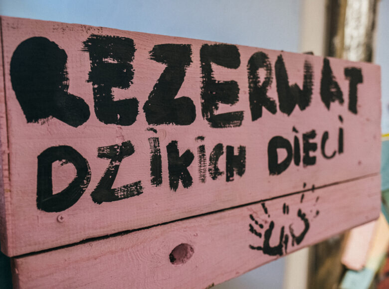 Zdajcie. Zbliżenie na drewnianą planszę. Plansza jest wykonana z desek, pomalowana na różowo. Czarną farbą namalowano napis: Rezerwat Dzikich Dzieci. Pod napisem odbite w czarnej farbie dwie dziecięce dłonie.