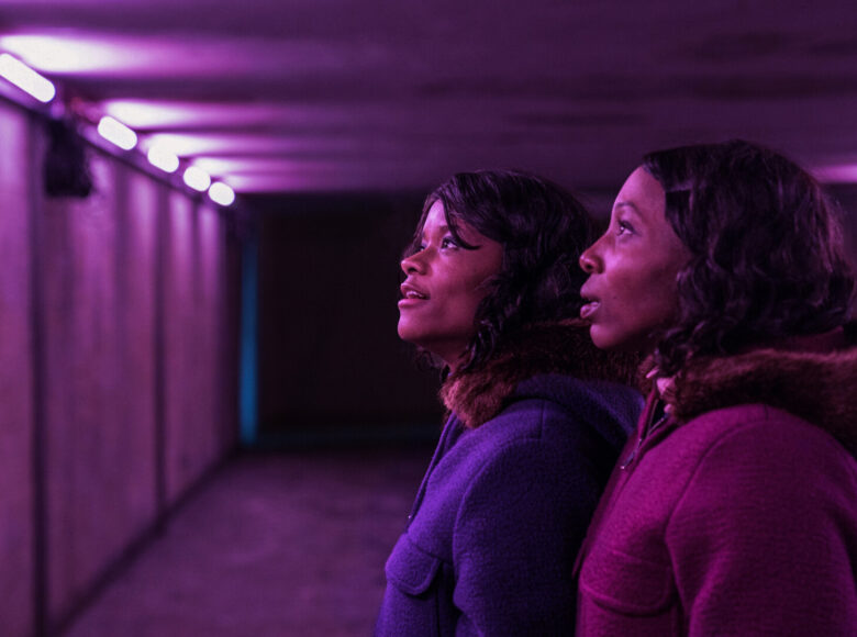 Dwie młode czarnoskóre kobiety, widziane z profilu, na zbliżeniu. Patrzą przed siebie, są oświetlone fioletowym światłem.