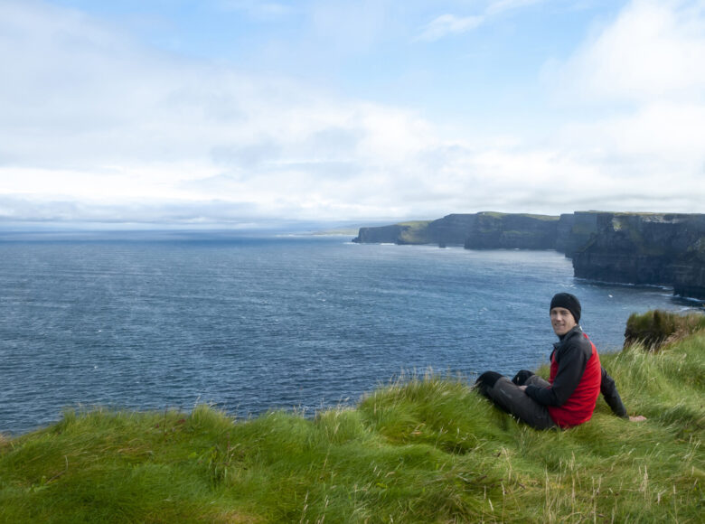 Zdjęcie krajobrazu. Widok z klifu. Na pierwszym planie zielona trawa porastająca klif. Na jego brzegu siedzi uśmiechnięty chłopak ubrany w czarne spodnie i czerwono-czarną bluzę. Na głowie ma czarną czapkę. W tle ciemnoniebieska woda oraz widok na dalszą część klifów. Lekko zachmurzone niebo.