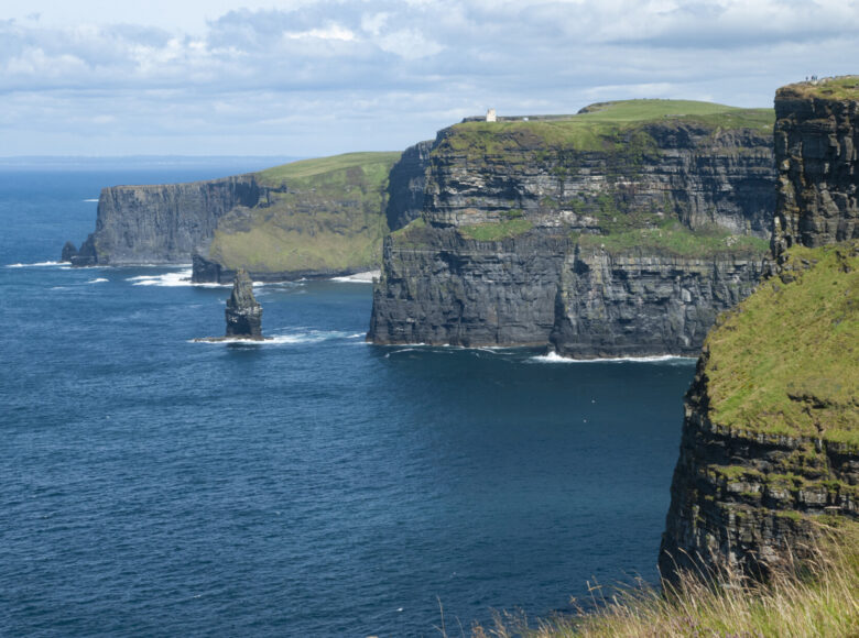 Zdjęcie krajobrazu. Po lewej stronie ciemnoniebieskie wody morza. W środkowej i prawej części kadru wysokie klify, które z wierzchu porasta intensywnie zielona roślinność. Na jednym z klifów widać malutkie sylwetki ludzi, na innym samotny budynek. Nad klifami lekko zachmurzone niebo.