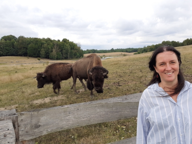 Zdjęcie przedstawia kobietę w jasnej koszuli w paski. Kobieta się uśmiecha, jej włosy rozwiewa wiatr. Za nią znajduje się ogrodzona drewnianym płotem łąka, na której stoją dwa żubry.