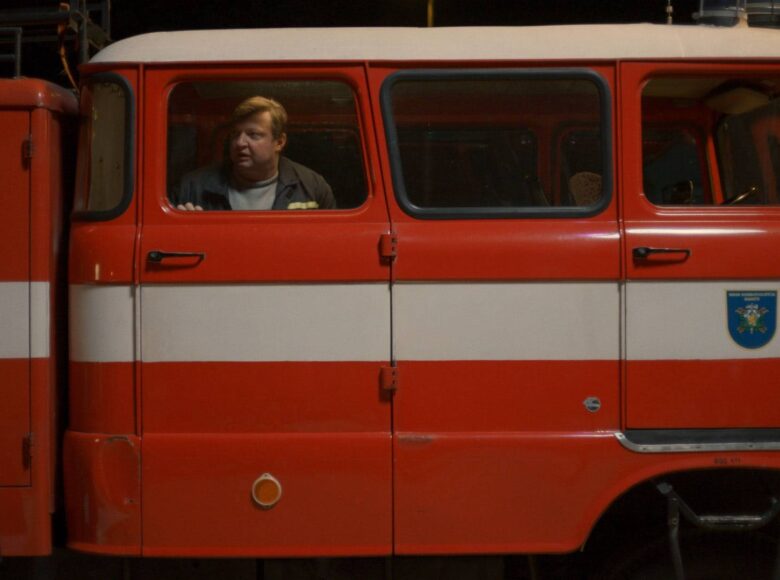 Kadr z filmu. Cały kadr wypełnia kabina pasażerska wozu strażackiego widziana z zewnątrz. Z wnętrza kabiny przez okno wygląda mężczyzna.