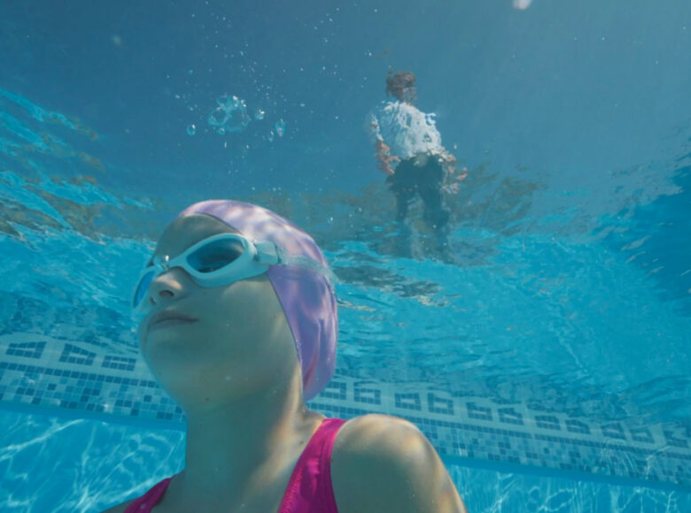 Kadr z filmu. Ujęcie spod wody. Na pierwszym planie głowa nurkującej dziewczynki w czepku i goglach pływackich, w tle zdeformowany obraz postaci stojącej nad powierzchnią wody