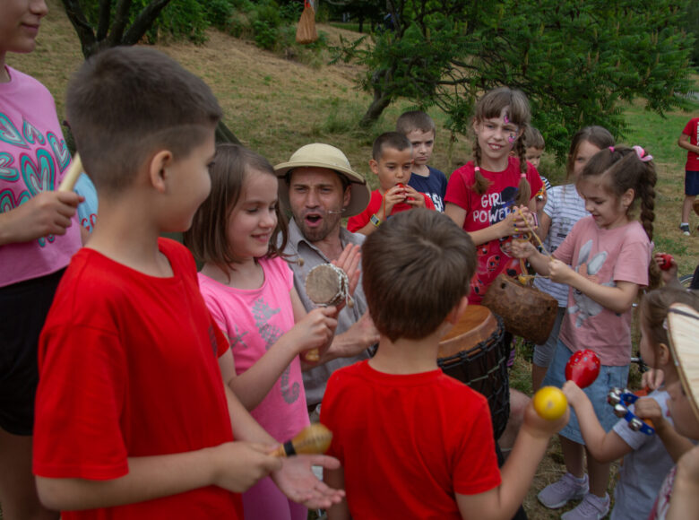 Zdjęcie. Scena ze spektaklu plenerowego. Mężczyzna w stroju podróżnika gra na bębnie. wkoło niego zgromadziły się dzieci grają na grzechotkach, bębenkach i cymbałkach.