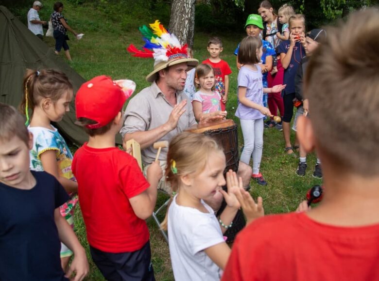 Zdjęcie. Przedstawia scenę ze spektaklu plenerowego pt. "Afryka Kazika". Mężczyzna w stroju podróżnika siedzi na krześle i gra na bębnie. Zgromadzone wokół niego dzieci klaszczą w dłonie.