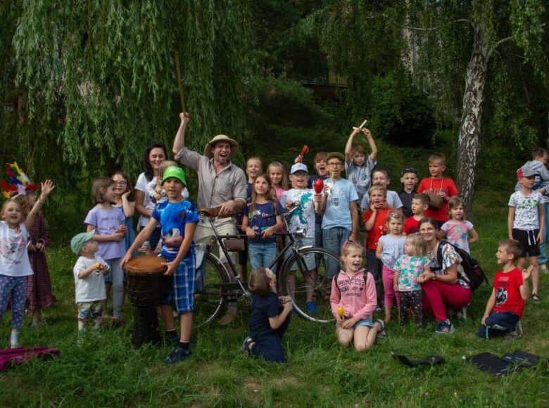 Zdjęcie. Scena z plenerowego spektaklu. Między drzewami soi mężczyzna, który trzyma przed sobą rower. Wokół mężczyzny zgromadzone są dzieci, które pozują do zdjęcia.