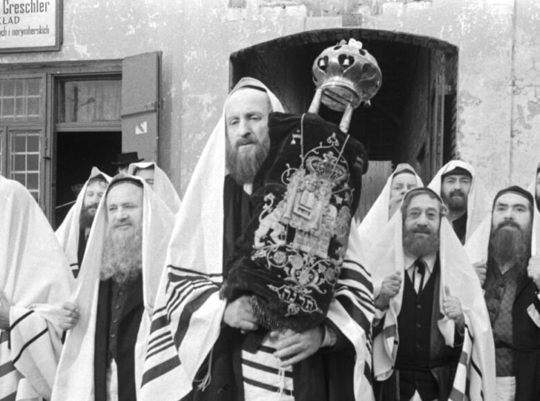 Kadr z czarno-białego filmu. Grupa religijnych Żydów stoi przed budynkiem. Wszyscy mają głowy nakryte tałesami – białymi chustami, a stojący na ich czele trzyma Torę w ozdobnym, haftowanym pokrowcu i z koroną.