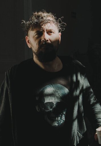 Zdjęcie. Portret mężczyzny w średnim wieku. Ma na sobie podkoszulek i ciemną bluzę. Na głowie nosi opaskę z drutu.