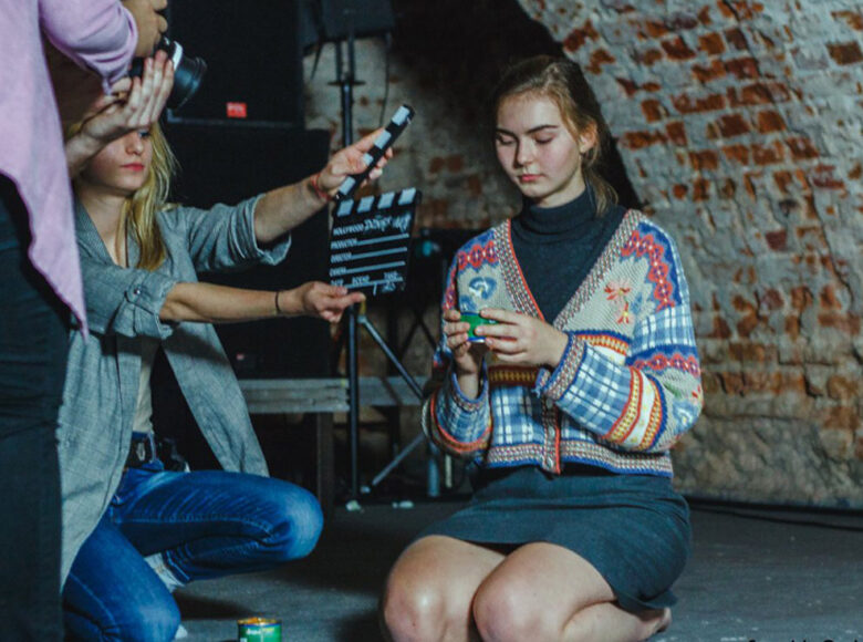 Zdjęcie. Trzy młode dziewczyny we wnętrzu. Jedna klęczy na podłodze, druga trzyma przed nią klaps filmowy, trzecia stoi przed obydwoma z aparatem fotograficznym w rękach.