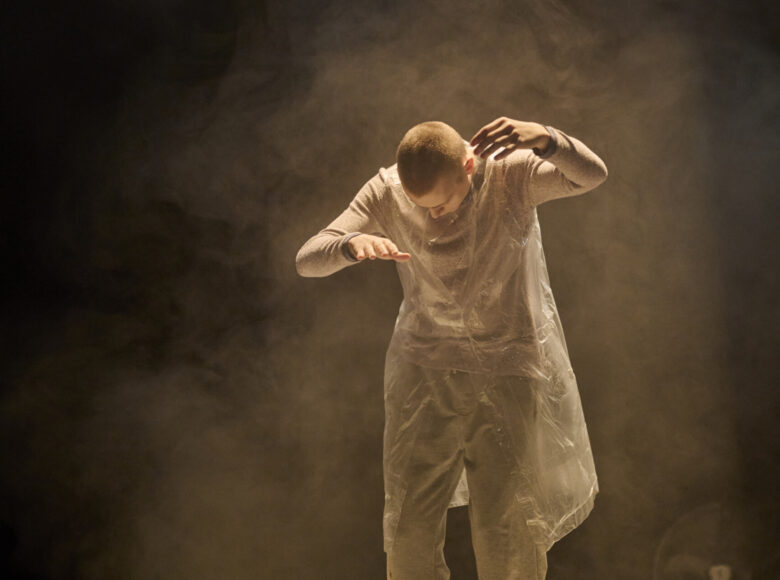 Zdjęcie przedstawia scenę ze spektaklu. Mężczyzna ubrany w szare spodnie, podkoszulek z długim rękawem i kamizelkę z folii unosi ręce przed siebie jakby kierował marionetką, patrzy w dół. Wokół niego unosi się dym.