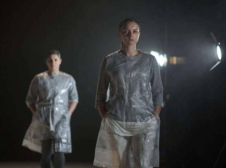 Zdjęcie ze spektaklu przedstawia dwie kobiety w szarych strojach i kamizelkach z przezroczystej folii.