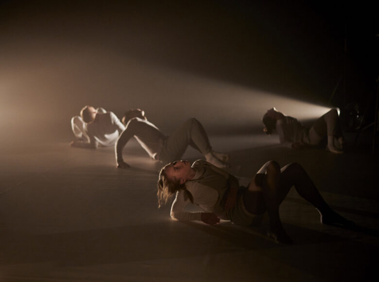 Zdjęcie przedstawia scenę ze spektaklu. Tancerze w szarych strojach siedzą na podłodze i patrzą w górę. Na scenę pada pojedynczy słup światła.