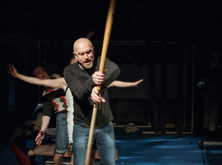 Kolorowe zdjęcie przedstawiające łysego mężczyznę w średnim wieku, który stoi na ciemnej scenie, pada na niego światło z góry. W dłoni trzyma długi drewniany kij przypominający wiosło. W tle widać dwie inne postaci: młodego chłopaka oraz dziewczyny, którzy stoją gęsiego za nim.