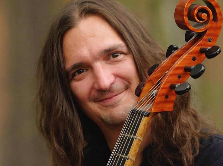 Zdjęcie. Przedstawia mężczyznę z długimi włosami, który trzyma wiolonczelę. Mężczyzna się uśmiecha.