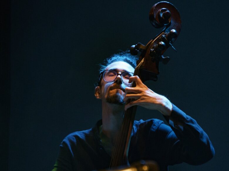 Zdjęcie. Fotografia przedstawia mężczyznę, muzyka, który gra na kontrabasie. Mężczyzna nosi okulary, ma zamknięte oczy, jego mina wyraża skupienie. Scena jest ciemna, na muzyka pada niebieskie światło.