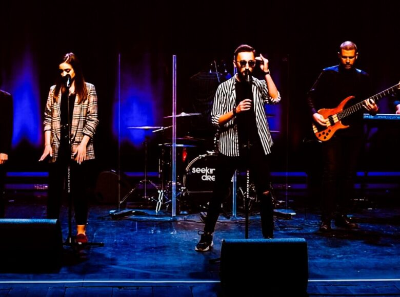 Zdjęcie. Na scenie oświetlonej niebieskimi lampami stoi zespół muzyczny. Przy mikrofonach stoją dwie kobiety i mężczyzna w ciemnych okularach. Na scenie znajdują się jeszcze trzej gitarzyści, perkusista i klawiszowiec.