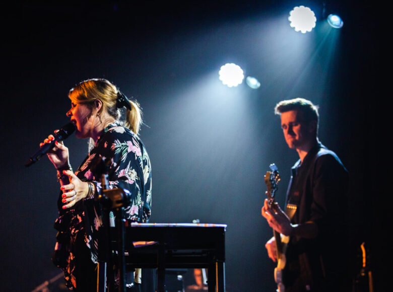 Zdjęcie przedstawia muzyków podczas koncertu. Kobieta śpiewa, mężczyzna, który stoi za nią gra na gitarze. Scena jest oświetlona pojedynczym snopem światła.