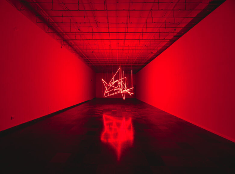 Zdjęcie. Wnętrze galerii sztuki. W pomieszczeniu znajduje się wystawa typu site-specific. Konstrukcja składa się z neonowych rur z których wydobywa się czerwone światło. Konstrukcja w oddaleniu odbija się w podłodze. Do sufitu pomieszczenia przymocowane są metalowe rury ułożone w kratownicę.