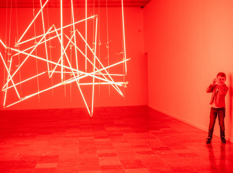 Zdjęcie. Wnętrze galerii sztuki. W pomieszczeniu znajduje się wystawa typu site-specific. Konstrukcja składa się z neonowych rur z których wydobywa się czerwone światło.. W pomieszczeniu znajduje się mały chłopiec, który robi zdjęcie instalacji.