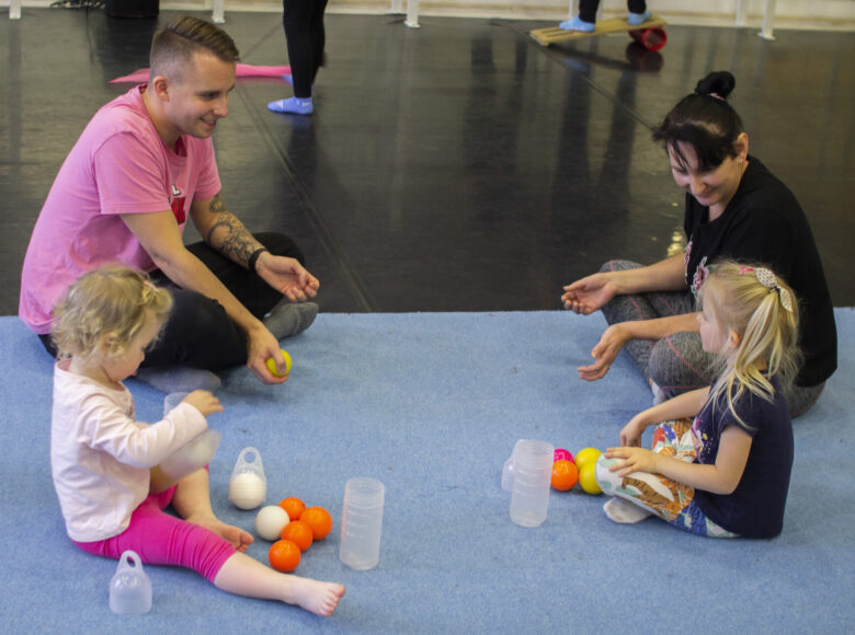 Zdjęcie. Na niebieskiej macie siedzi para dorosłych: kobieta i mężczyzna oraz dwie małe dziewczynki. Obok nich lezą żółte piłeczki, mandarynki i pławikowe kubeczki.