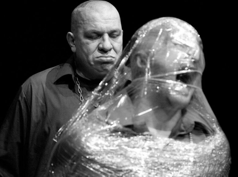 Czarno-białe zdjęcie. Umięśniony mężczyzna z łańcuchem na szyi stoi z groźną miną i patrzy w dół. Przed nim stoi mężczyzna owinięty przezroczystą folią jak kokonem. Jego twarz wyraża przerażenie.