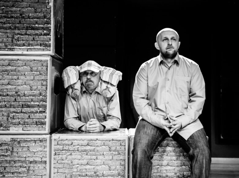 Czarno-białe zdjęcie. Scena ze spektaklu "Zły". Dwóch mężczyzn siedzi na ceglanym murku. Jeden z nich ma na głowie czapkę z daszkiem z przyklejonymi do niej dwoma papierowymi twarzami.