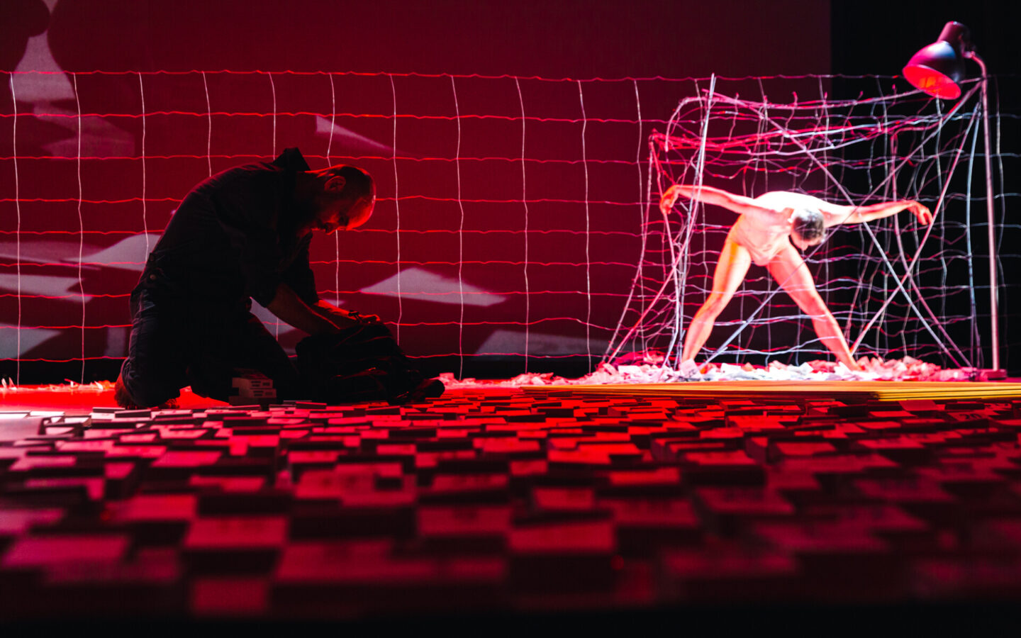 Zdjęcie przedstawia scenę ze spektaklu. Podłoga jest usłana kartkami z kalendarza, wśród nich klęczy mężczyzna i pakuje plecak. Na drugim planie znajduje się zapora i klatka z drutu kolczastego. W klatce znajduje się półnagi mężczyzna. Stoi w szerokim rozkroku ma rozłożone w bok ręce i pochyla się do przodu. Na scenę pada czerwone światło.