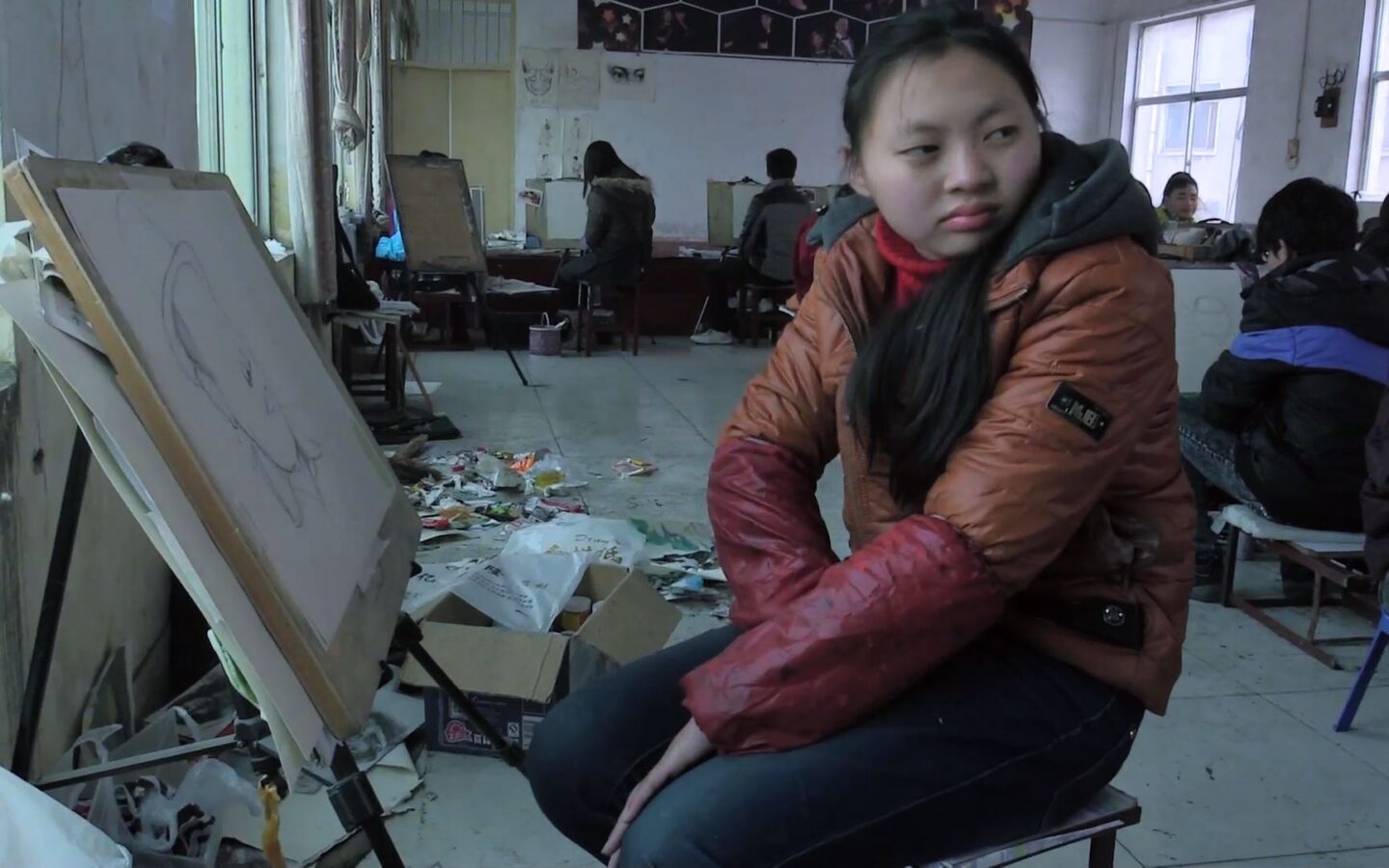 Kadr z filmu. Wnętrze sali szkolnej. Na pierwszym planie dziewczynka, azjatka, siedzi przed sztalugą. Jest ubrana w kurtkę i jeansy. Wokoło inne dzieci przed sztalugami, Pomieszczenie jest zaniedbane.