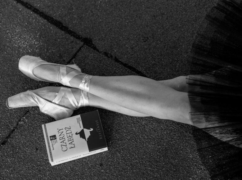 Czarno-białe zdjęcie przedstawia nogi kobiety w butach do tańczenia baletu. Na ziemi leży książka pt. "Czarny łabędź".