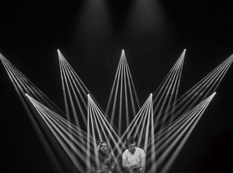 Na podłodze ciemnej sceny siedzi dwóch młodych mężczyzn. Jeden z nich ma krótkie włosy i jasną koszulkę, drugi jest ubrany na ciemno, ma brodę i włosy do ramion. Mężczyzn oświetla punktowe, krzyżujące się na scenie światło reflektorów płynące z wielu stron.
