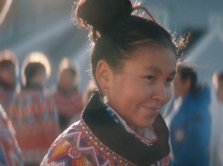 Kadr z filmu. Zbliżenie na młodą dziewczyną o ciemnych włosach upiętych w kok i o skośnych oczach. Ubrana w kolorową, wzorzystą odzież. Tło nieostre, ale widać tam inne osoby w podobnych ubraniach.