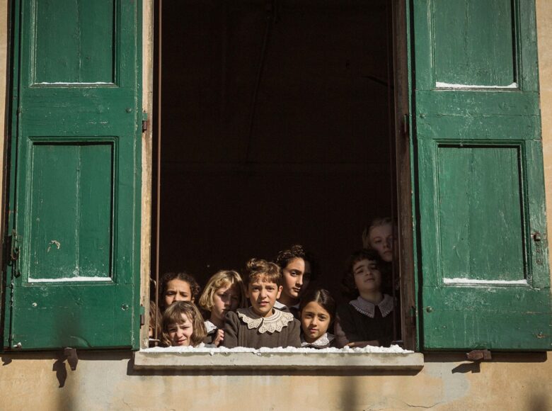 Kadr z filmu. Grupa 9 dzieczynek stojących w otwartym oknie, widziana z zewnątrz. Dziewczynki są ubrane w szare sukienki z białymi kołnierzykami. Przy oknie, otwarte na oścież zielone okiennice.