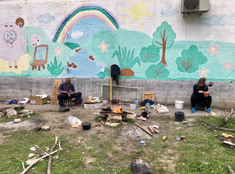 Zdjęcie przedstawia podwórko. Na murach budynku namalowano mural, który przedstawia tęcze, łakę, rzekę, słońce, kwiaty i baranka, który maluje obraz. Pod ścianą siedzi dwoje ludzi: męzczyzna i kobieta. Są bezdomni. Przed nimi w nieładzie leżą różne przedmioty: kuki, garnki, kawałki drewna, kartony, butelki z wodą.