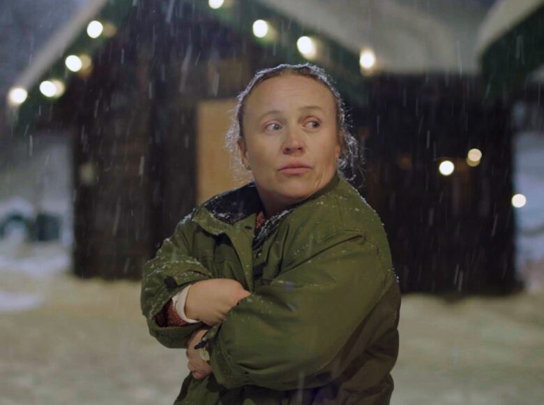 Kadr z filmu. Zbliżenie na niskorosłą kobietę stojącą na zewnątrz. Kobieta jest w zielonej kurtce, w rozmytym tle widać ośnieżone domki ze światełkami i padający śnieg. Kobieta ma założone ręce, stoi na wprost nas, ale patrzy w bok.