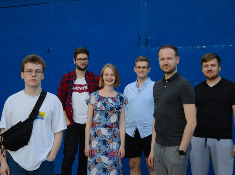 Na tle granatowych metalowych drzwi stoi grupa młodych ludzi: jedna kobieta i pięciu mężczyzn. Mają na sobie lekkie, wiosenno-letnie stroje, patrzą z uśmiechem w obiektyw.