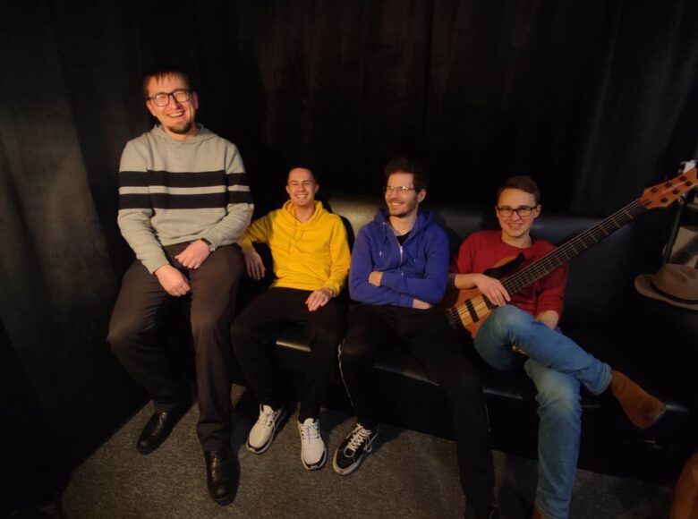 Na tle ciemnej ściany na ciemnej kanapie siedzi czterech młodych mężczyzn w casualowych strojach. Są w pogodnych nastrojach, jeden z nich trzyma w ręku gitarę.