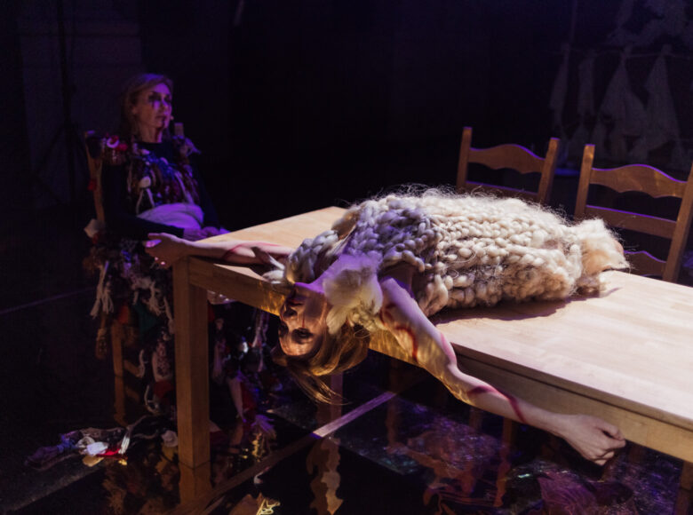 Zdjęcie przedstawia scenę ze spektaklu "Dziady". Na scenę pada fioletowe światło. Na drewnianym stole leży dziewczyna w kostiume zrobionym z jasnych, splecionych w warkocz włókien.