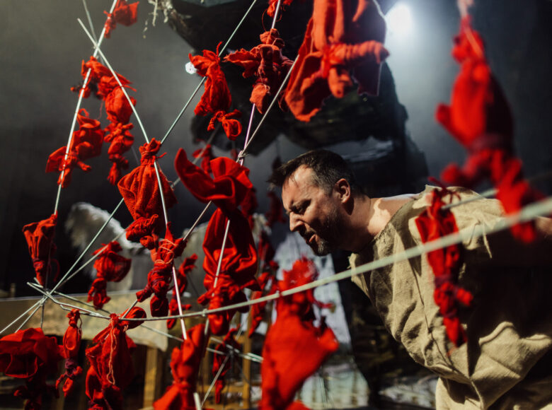 Zdjęcie przedstawia scenę ze spektaklu "Dziady". Mężczyzna w tunice przypominającej jutowy worek znajduje się w klatce wykonanej z drutu i czerwonych lalek motanek.