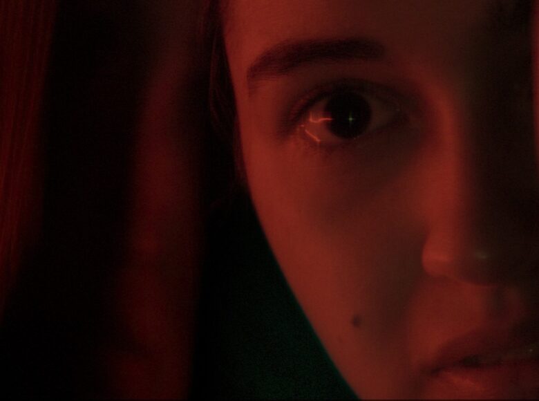 Zbliżenie na połowę twarzy młodej dziewczyny. Twarz i rozmyte tło są czerwone. Widać jej szeroko otwarte oko i lekko rozchylone usta.