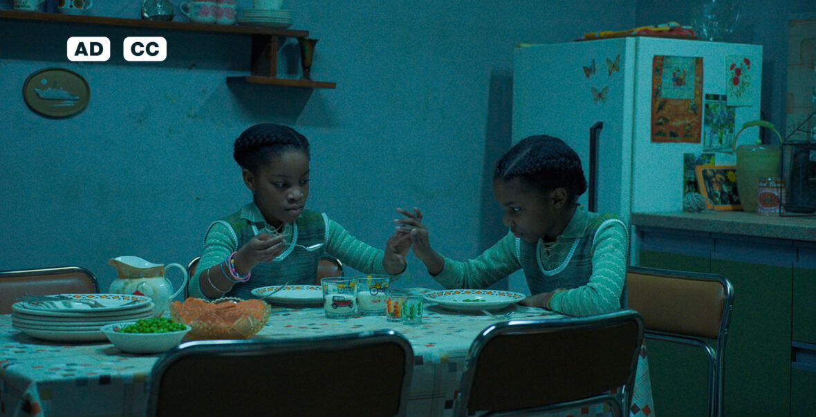 Na zdjęciu dwie czarnoskóre dziewczynki. Siedzą za stołem w kuchni, przed nimi talerze. Trzymają się za ręce.